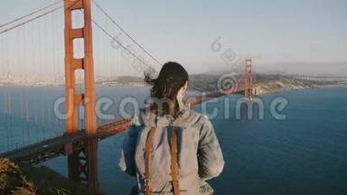 后景相机放大年轻女子背包，头发吹在风中欣赏日落在金门大桥。
